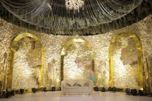 A luxurious wedding setting with a floral arch adorned with vibrant flowers, leading to a beautifully decorated stage with cascading floral chandeliers and intricate floral arrangements.