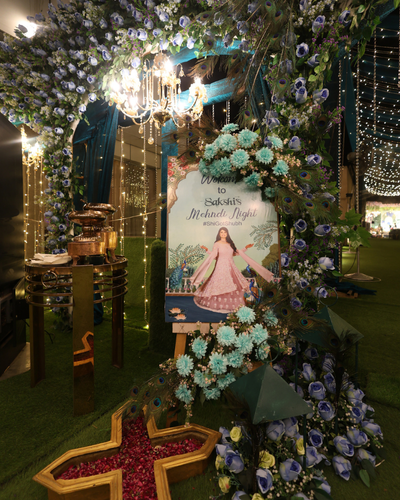 Decorative entrance arch with floral arrangement and welcome board for Sakshi's Mehndi Night celebration.