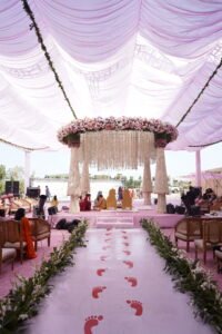 An outdoor wedding ceremony with a pink floral mandap, a path marked with red footprints, and guests seated in chairs, all under a large, draped canopy.