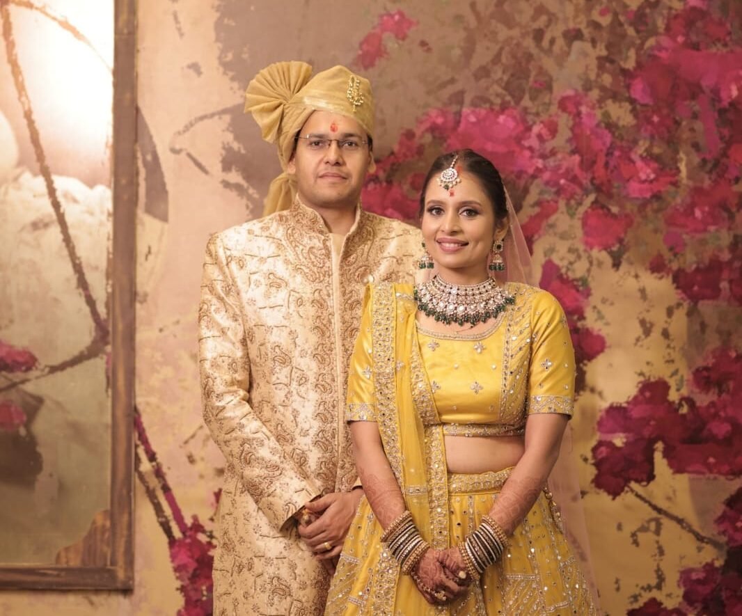 Bride and groom in traditional golden and yellow Indian wedding attire posing in front of a floral backdrop.