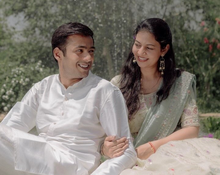 Couple sitting outdoors in traditional Indian attire, smiling at each other with a soft natural background.