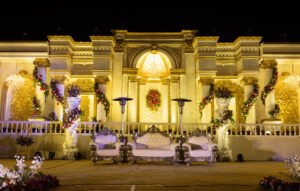 A grand wedding stage with an opulent golden backdrop, adorned with cascading floral arrangements, a chandelier, and luxurious white and silver seating for the couple.
