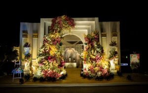 A grand floral wedding entrance with towering flower arrangements in pink, white, and yellow hues, illuminated by soft lighting, leading to an ornate backdrop with elegant columns.