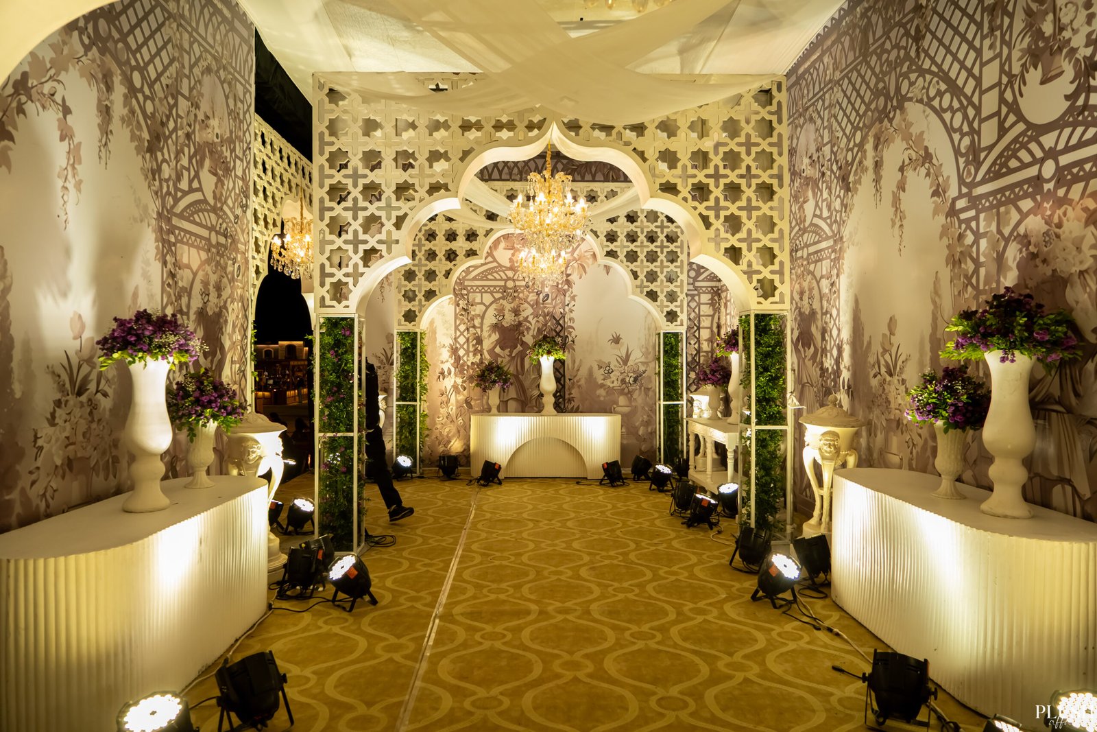 A beautifully designed wedding stage with intricate white arches, floral wallpaper, and elegant floral arrangements, illuminated by soft lighting and a central chandelier.