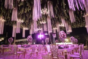 A glamorous wedding reception setup featuring elegant chandeliers and hanging floral strands above luxurious tables adorned with vibrant floral arrangements, illuminated by soft purple lighting.