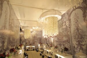 A luxurious wedding venue featuring chandeliers made of cascading flowers, a floral wallpaper backdrop, and elegant white tables adorned with colorful flower arrangements.