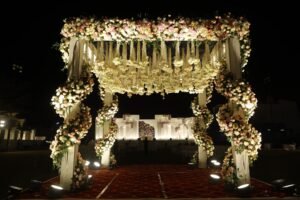 A stunning outdoor wedding mandap draped with cascading white and pink flowers, illuminated by soft lighting, creating a romantic and enchanting atmosphere under the night sky.