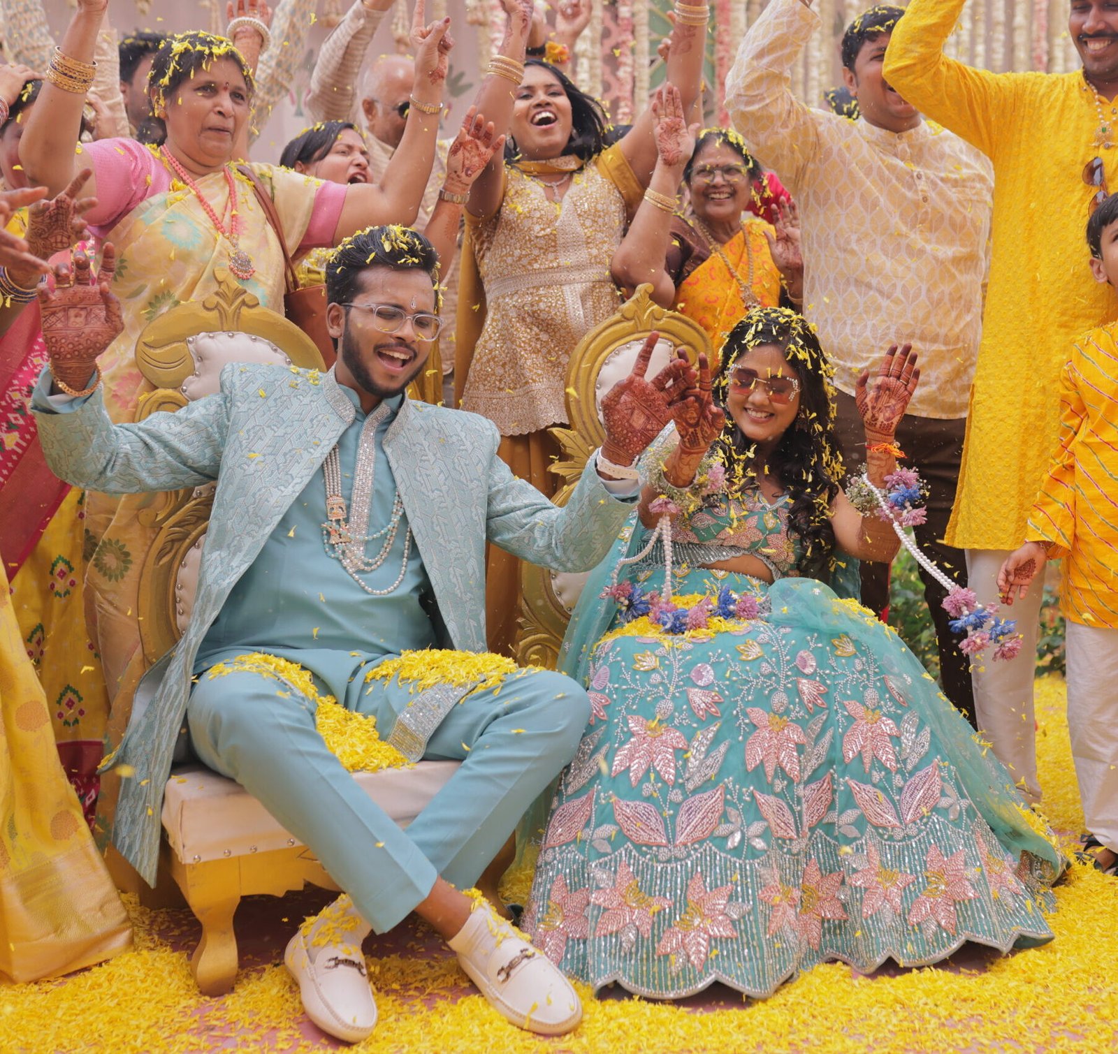 The bride and groom enjoy a joyous moment during their wedding ceremony, surrounded by family members who cheer and celebrate. The couple, seated on golden chairs, is showered with yellow flower petals, adding a festive touch to the scene. Everyone is smiling and laughing as they celebrate the special occasion.