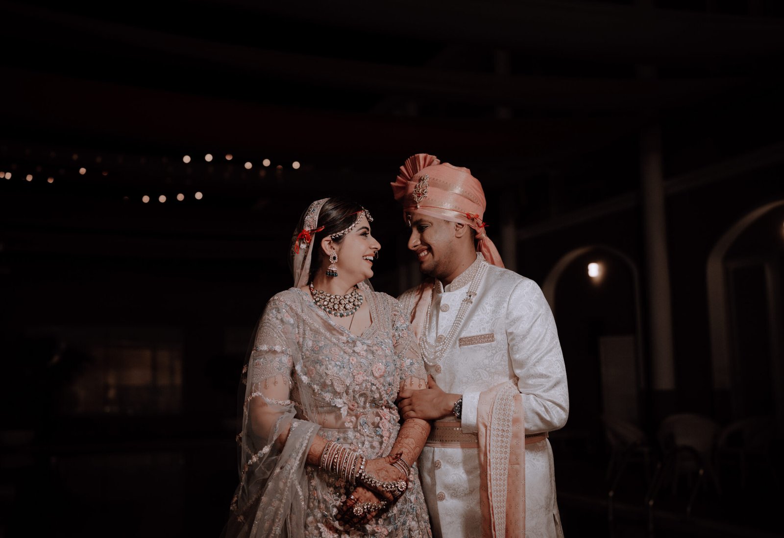 Bride and groom in pastel wedding attire smiling at each other during an intimate indoor moment.