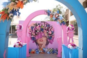 A colorful wedding entrance featuring a vibrant pink and blue archway with floral arrangements, leading to a whimsical art piece of a woman’s face adorned with flowers and pink sculptures.
