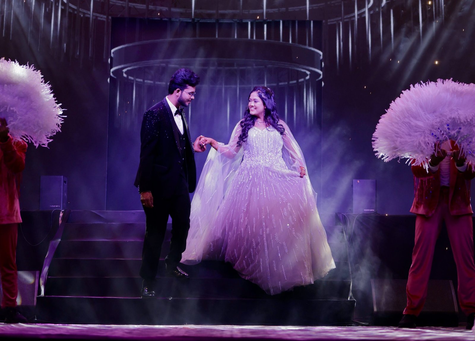Couple making a grand entry on stage with spotlight and sparkling gown in a romantic setting.