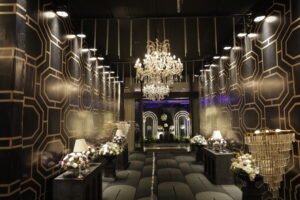 A luxurious wedding venue with a modern geometric-patterned wall, elegant chandeliers, and floral arrangements along the aisle, creating a sophisticated and glamorous atmosphere.