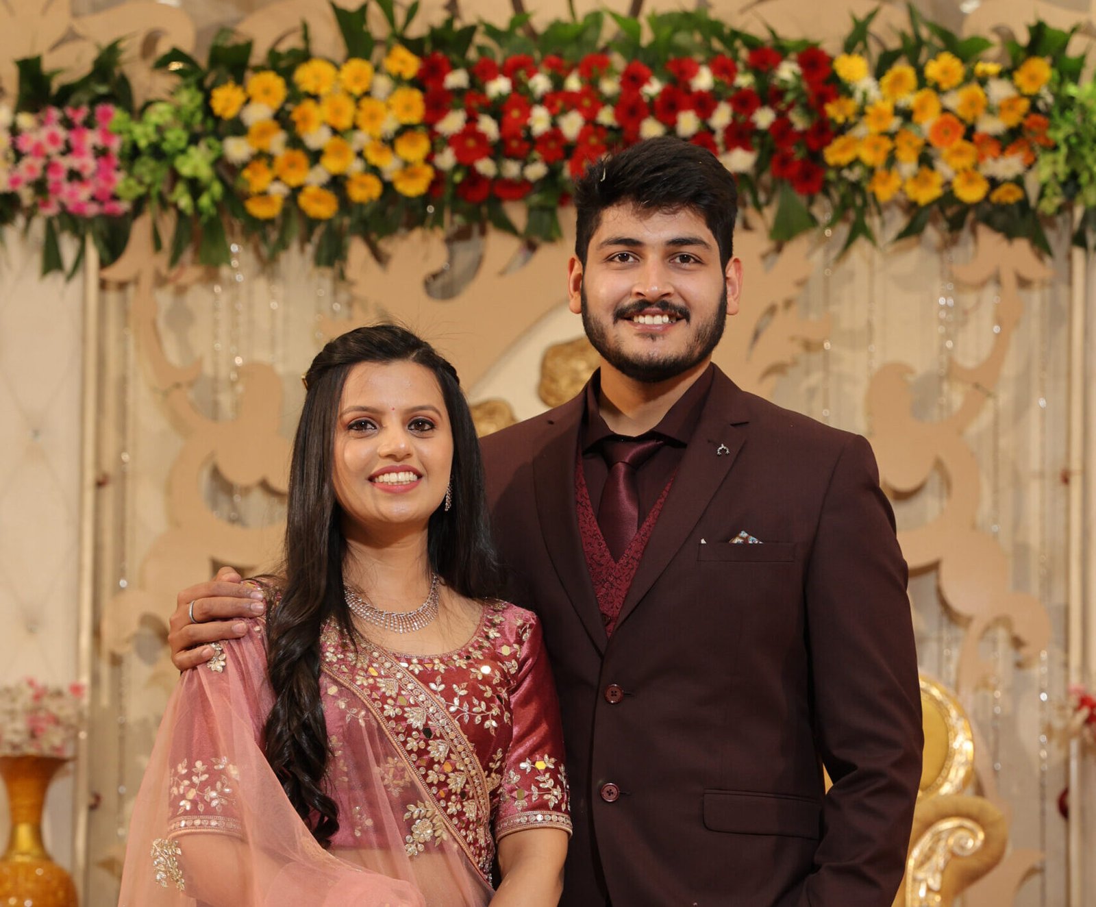 Couple smiling at their engagement ceremony in traditional Indian attire with floral backdrop.