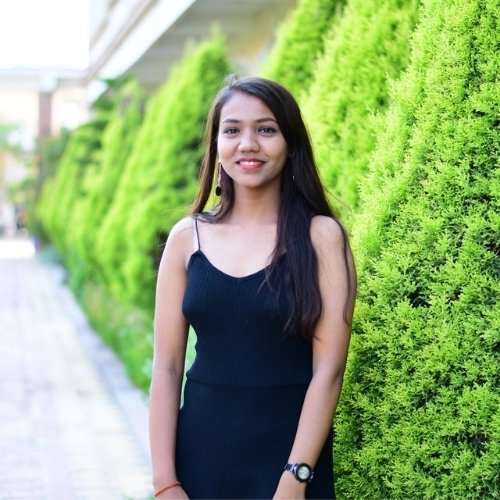 Anamika Kushwaha smiling confidently in a black dress near vibrant green hedges