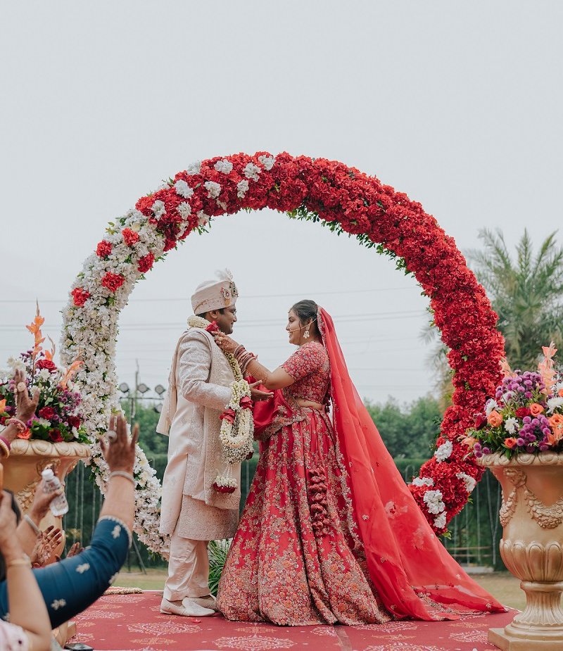 Indian bride and groom exchanging garlands under floral arch during outdoor wedding ceremony